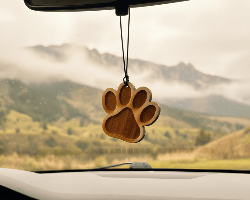 Wooden paw-shaped car air freshener hanging from a car's rearview mirror with a scenic view outside.
