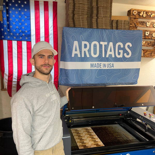 Person standing in front of a large American flag and a laser engraving cutting machine with Arotags' branding.